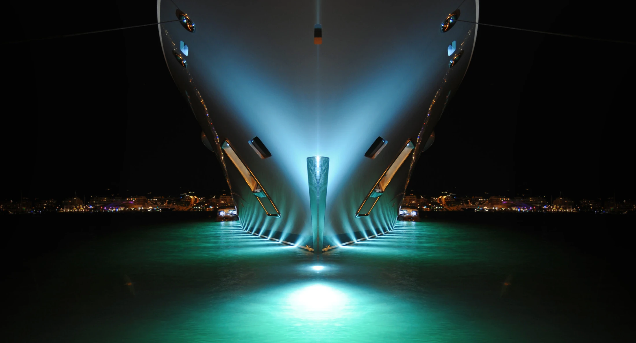 The bow of a superyacht illuminated at night above turquoise water